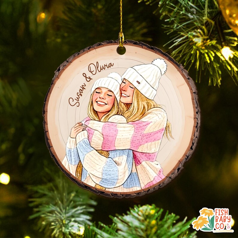 May include: A round wooden Christmas ornament with a drawing of two smiling people hugging, wearing white knit hats and striped sweaters. The ornament has the names "Susan & Olivia" written on it. The ornament is hanging from a gold string.