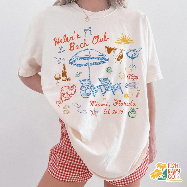 May include: Cream-colored t-shirt with a beach-themed graphic. The design includes an umbrella, beach chairs, and the text "Helen's Bach Club Miami, Florida Est. 2025". The shirt is paired with red and white checkered shorts.