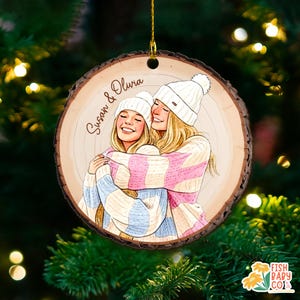 May include: A wooden Christmas ornament with a drawing of two women hugging, wearing white beanies and striped sweaters in pink, blue, and cream. The ornament has the names "Susan & Olivia" written on it.