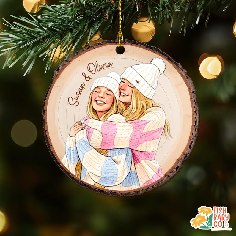 May include: A wooden Christmas ornament with a drawing of two women hugging, wearing white beanies and striped sweaters in pink and blue. The ornament has the names "Susan & Olivia" written on it. The ornament is hanging from a gold string.
