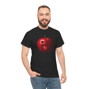 May include: Black t-shirt featuring a red apple with a face and a tongue sticking out. The apple has eyes and a stem. The t-shirt is made of a soft material and is suitable for casual wear.