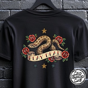 Puede incluir: Camiseta negra con un diseño de tatuaje. Presenta una serpiente, rosas rojas y estrellas. Una pancarta en el diseño dice "PLAY DEAD". La camiseta tiene cuello redondo. El diseño está centrado.