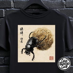 May include: Black t-shirt with a detailed illustration of a beetle pushing a ball of dirt, Japanese calligraphy, and a red seal. The design is in a traditional style with a vintage aesthetic. The shirt also has a Cold Blooded Club logo.