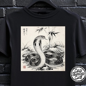 May include: Black t-shirt featuring a detailed black and white illustration of a cobra with bamboo in the background. The snake is coiled, with its tongue extended. The shirt has a round logo on the bottom right.