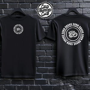 May include: Two black t-shirts are displayed, one showing the front and the other the back. Each shirt features a white circular design with the text "CORN SNAKE" and a snake illustration. The shirts are hanging against a dark brick wall.