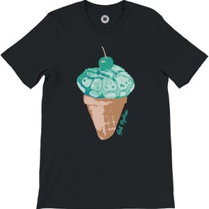 K&ouml;nnte beinhalten: Schwarzes T-Shirt mit einer skurrilen Illustration eines eiscreme-f&ouml;rmigen Eis mit Schlangenmuster, gekr&ouml;nt von einer Kirsche. Der Kegel ist braun und das Eis ist t&uuml;rkis. Der Text "Ball Python" ist t&uuml;rkis auf dem Kegel geschrieben.