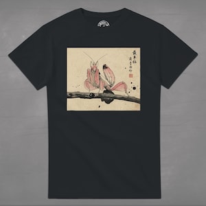 May include: Black t-shirt featuring a detailed illustration of a praying mantis in shades of pink and brown, perched on a branch. The artwork is set against a beige background with Japanese text and ink splatters, creating a unique design.