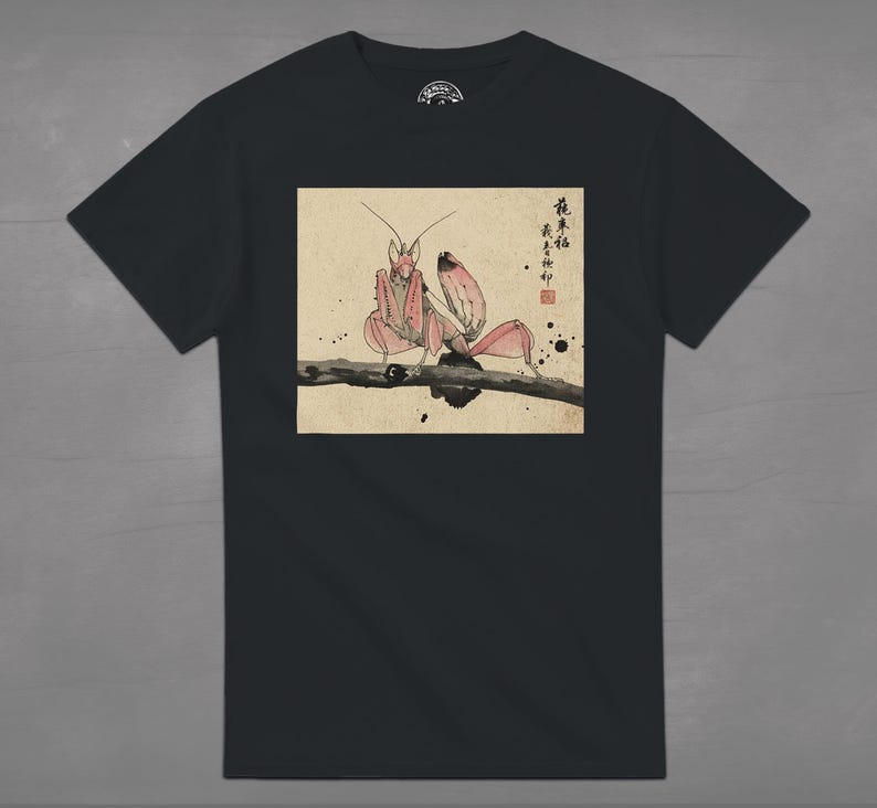 May include: Black t-shirt featuring a detailed illustration of a praying mantis in shades of pink and brown, perched on a branch. The artwork is set against a beige background with Japanese text and ink splatters, creating a unique design.