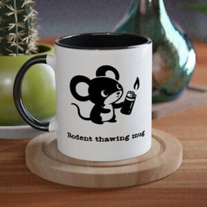 Puede incluir: Taza de cerámica blanca con interior y asa negros. La taza presenta un dibujo de un ratón negro de dibujos animados sosteniendo un encendedor, con el texto "Rodent thawing mug" debajo de la imagen. La taza está sobre un posavasos de madera.
