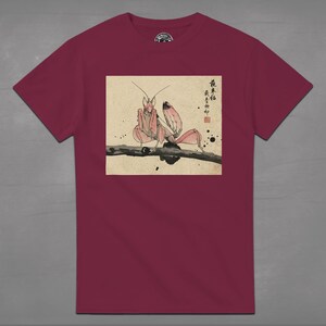 May include: A maroon t-shirt featuring a detailed illustration of a praying mantis perched on a branch. The artwork is in a traditional Japanese style, with soft pink and brown tones. The shirt has a crew neck and short sleeves.