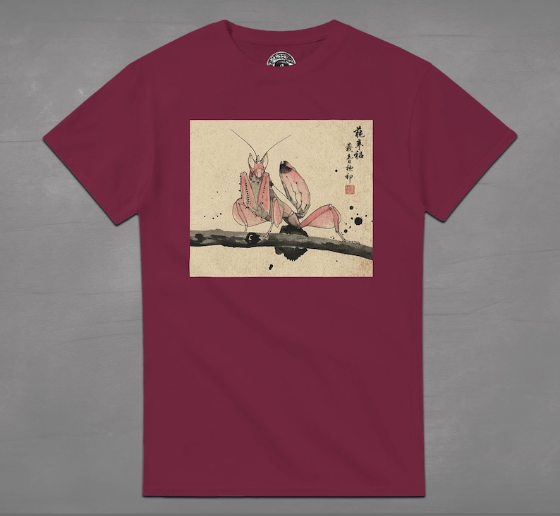 May include: A maroon t-shirt featuring a detailed illustration of a praying mantis perched on a branch. The artwork is in a traditional Japanese style, with soft pink and brown tones. The shirt has a crew neck and short sleeves.