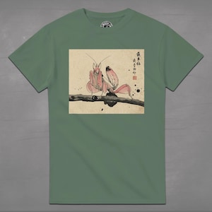 May include: Sage green t-shirt featuring a detailed illustration of a pink praying mantis perched on a branch. The artwork is in a traditional Japanese style, with calligraphy and ink blotches. The shirt has a crew neck and short sleeves.
