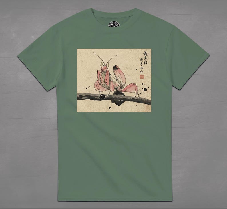 May include: Sage green t-shirt featuring a detailed illustration of a pink praying mantis perched on a branch. The artwork is in a traditional Japanese style, with calligraphy and ink blotches. The shirt has a crew neck and short sleeves.