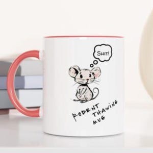 Puede incluir: Taza de cerámica blanca con interior y asa rojos. La taza presenta una ilustración de ratón de dibujos animados con la palabra "Shit!" en un bocadillo. El texto "Rodent Thawing Mug" está debajo del ratón.