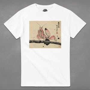 May include: White t-shirt featuring a pink praying mantis illustration in a Japanese art style. The insect is perched on a branch, with Japanese characters and ink splatters in the background. The shirt has a crew neck and short sleeves.