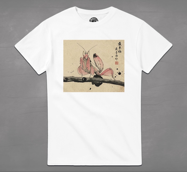 May include: White t-shirt featuring a pink praying mantis illustration in a Japanese art style. The insect is perched on a branch, with Japanese characters and ink splatters in the background. The shirt has a crew neck and short sleeves.