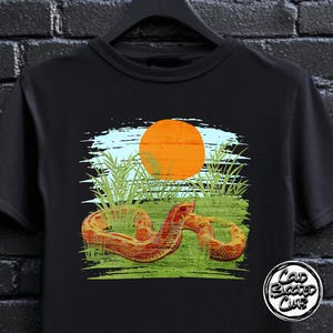 Puede incluir: Camiseta negra con un gráfico de una serpiente en un campo de hierba con un sol naranja. La serpiente es roja y marrón, y la hierba es verde. La camiseta tiene el texto "Cold Blooded Club" en la parte inferior derecha.
