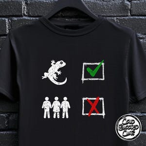 Op de afbeelding: Zwart T-shirt met een witte gekko-afbeelding, een groen vinkje en een rode X. Het shirt heeft ook de tekst "Cold Blooded Club" in een cirkelvormig ontwerp.