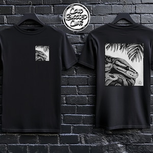 May include: Two black t-shirts featuring a snake graphic. One shirt has a small square image on the front, the other a larger image on the back. The graphic is a detailed black and white drawing of a snake with palm leaves. The shirts are hanging on black hangers.