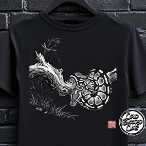 Pode incluir: Camiseta preta com uma ilustração em preto e branco de uma cobra enrolada em um galho, com bambu. O design inclui o texto "Cold Blooded Club" em um logotipo circular e um selo japonês.