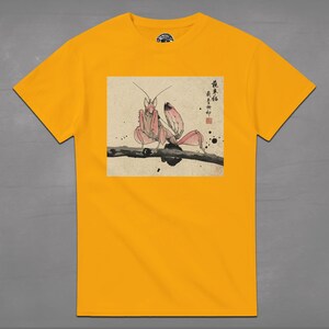 May include: Mustard yellow t-shirt featuring a detailed illustration of a pink praying mantis perched on a branch. The artwork is in a traditional Japanese style, with calligraphy and ink blotches. The shirt is a classic crew neck.