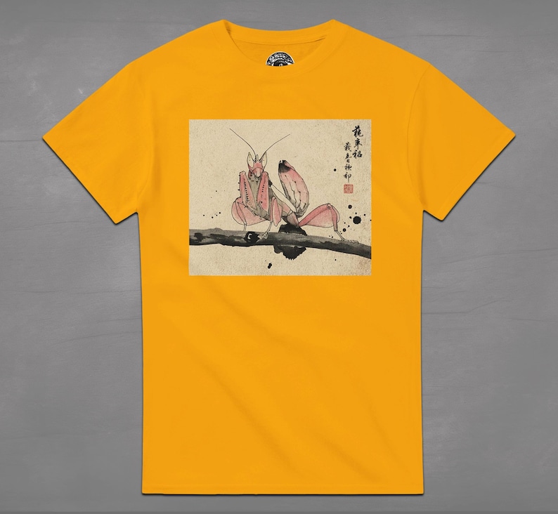 May include: Mustard yellow t-shirt featuring a detailed illustration of a pink praying mantis perched on a branch. The artwork is in a traditional Japanese style, with calligraphy and ink blotches. The shirt is a classic crew neck.