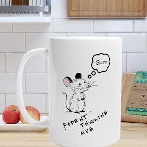 Może przedstawiać: Biały ceramiczny kubek z ilustracją myszy z kreskówki i napisem "Rodent Thawing Mug". Mysz ma dymek z napisem "Shit!". Dwa czerwone jabłka leżą przed kubkiem.