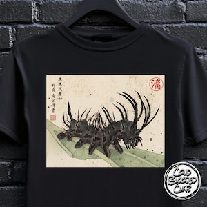 May include: Black t-shirt featuring a detailed illustration of a spiky, dark-colored insect-like creature on a leaf. The design has a vintage, artistic style with Japanese text and a red seal. The shirt also has a "Cold Blooded Club" logo.