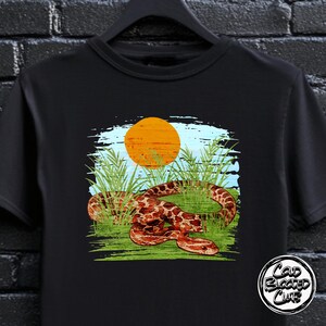 May include: Black t-shirt with a graphic of a snake in a grassy field with a setting sun. The snake is brown with a patterned design. The design includes the text "Cold Blooded Club" in a circular logo.