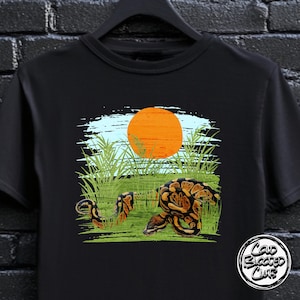 Peut inclure: T-shirt noir avec un motif de serpent dans un champ herbeux, un soleil orange et un ciel bleu. Le design comprend le texte "Cold Blooded Club" dans un logo circulaire en bas à droite.