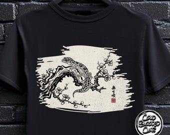 Może przedstawiać: Czarny t-shirt z czarno-białą ilustracją gekona na kwitnącej gałęzi. Projekt zawiera japoński tekst i czerwoną pieczęć. Okrągłe logo z napisem "Cold Blooded Club" znajduje się w prawym dolnym rogu.