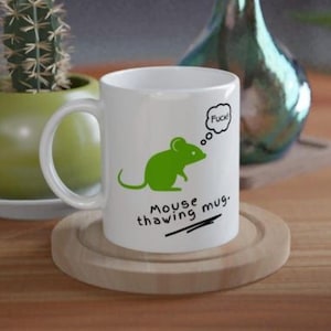 May include: White ceramic mug with a green mouse graphic and the text "Mouse thawing mug." A thought bubble above the mouse says "Fuck!" The mug sits on a wooden coaster.