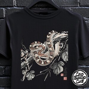 May include: Black t-shirt with a detailed illustration of a snake coiled around a branch, with leaves and Japanese text. The snake has brown, tan, and white markings. A circular logo with the text "Cold Blooded Club" is on the bottom right.