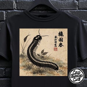 以下が含まれることがあります： 黒いTシャツ。日本の伝統的なスタイルで描かれたムカデのイラストが特徴です。 日本語の文字、赤い印鑑、草や葉のあるベージュ色の背景が含まれています。 白いロゴが右下にあります。