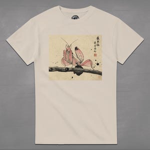 May include: Cream-colored t-shirt featuring a Japanese-style illustration of a praying mantis in shades of pink and red, perched on a branch. The artwork includes Japanese characters and ink splatters, creating a unique design.