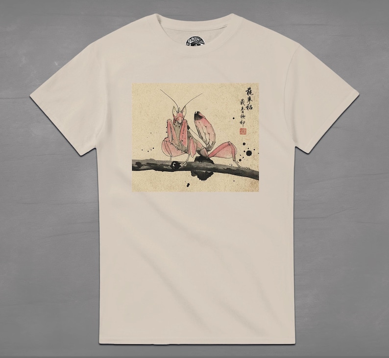 May include: Cream-colored t-shirt featuring a Japanese-style illustration of a praying mantis in shades of pink and red, perched on a branch. The artwork includes Japanese characters and ink splatters, creating a unique design.