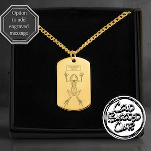 May include: A gold-coloured dog tag pendant with a chain. The tag features an engraved skeletal illustration of a frog and the words "COMMON FROG". The pendant is displayed in a black box with the text "Cold Blooded Club".