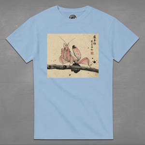 May include: Light blue t-shirt featuring a pink praying mantis illustration on a branch. The artwork has a Japanese aesthetic with calligraphy and ink blotches. The shirt is a short-sleeved crew neck.
