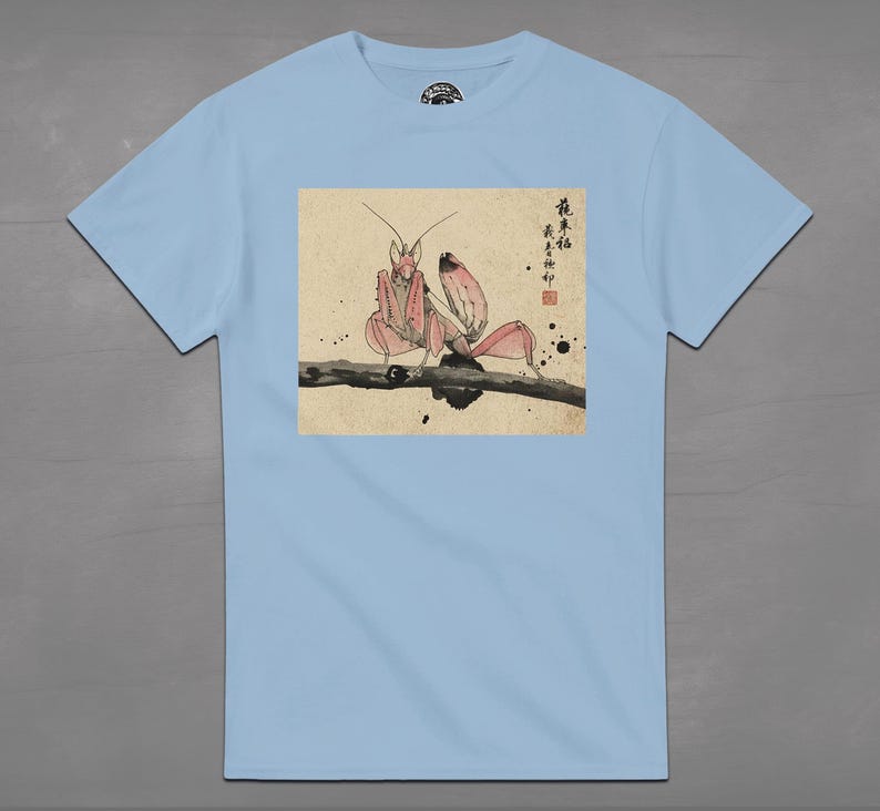 May include: Light blue t-shirt featuring a pink praying mantis illustration on a branch. The artwork has a Japanese aesthetic with calligraphy and ink blotches. The shirt is a short-sleeved crew neck.