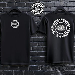 Peut inclure: Deux t-shirts noirs, présentés de face et de dos, chacun avec un motif circulaire blanc. Le motif avant indique "ROYAL PYTHON" autour d'une illustration de serpent. Le motif arrière répète le texte et l'illustration. Un logo "Cold Blooded Club" est visible.