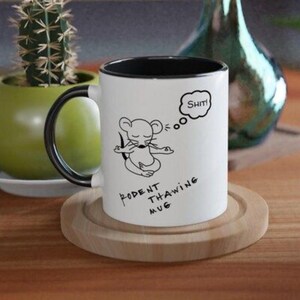 Puede incluir: Taza de cerámica blanca con interior y asa negros. La taza presenta un ratón de dibujos animados meditando con la palabra "Shit!" en una burbuja de pensamiento. El texto "RODENT THAWING MUG" también está impreso.