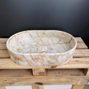 May include: Oval-shaped marble sink with a cream and gold veined pattern. The sink has a smooth, polished surface and a central drain. It is displayed on a wooden pallet, showcasing its natural stone texture and design.