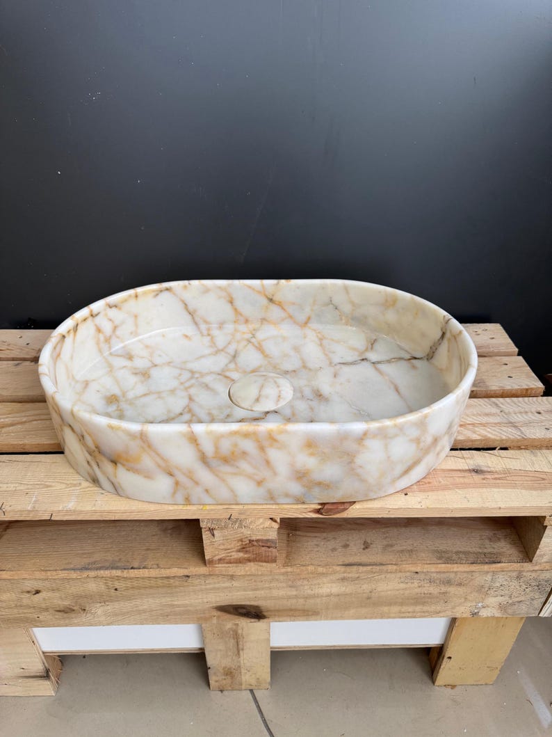 May include: Oval-shaped marble sink with a cream and gold veined pattern. The sink has a smooth, polished surface and a central drain. It is displayed on a wooden pallet, showcasing its natural stone texture and design.