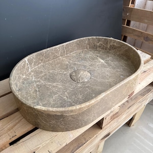 May include: Oval-shaped bathroom sink with a marbled, brown finish. The sink has a central drain and is set on a wooden pallet. The sink's design features a smooth, rounded interior and a textured exterior.