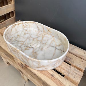 May include: Oval-shaped marble sink with a white and gold veined pattern. The sink has a central drain hole and sits on a wooden pallet. The sink's interior is smooth and polished, showcasing the natural stone's intricate design.