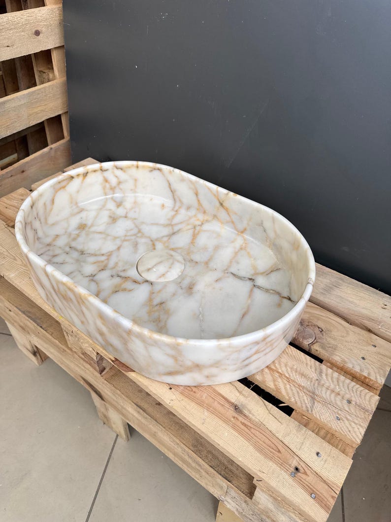 May include: Oval-shaped marble sink with a white and gold veined pattern. The sink has a central drain hole and sits on a wooden pallet. The sink's interior is smooth and polished, showcasing the natural stone's intricate design.