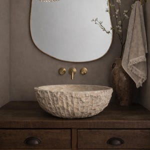 May include: A bathroom vanity with a textured stone sink, brass fixtures, and a large oval mirror with a gold frame. A vase with branches and a towel complete the neutral-toned, spa-like aesthetic.