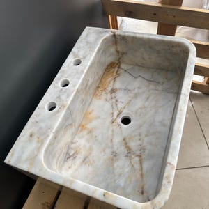 May include: A rectangular marble sink with a rounded basin and three tap holes. The sink features a white and beige veined pattern. The sink is set on a wooden pallet.