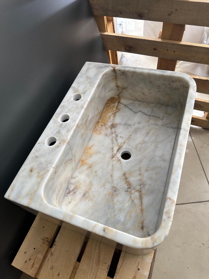 May include: A rectangular marble sink with a rounded basin and three tap holes. The sink features a white and beige veined pattern. The sink is set on a wooden pallet.