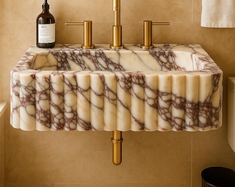 May include: A rectangular marble sink with a scalloped edge and a gold faucet. The sink has a cream base with burgundy veining. A matching gold towel ring and a black soap dispenser are also visible.